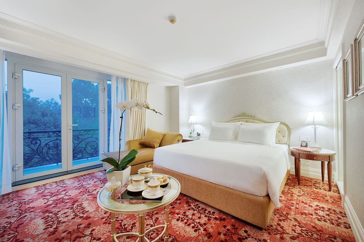 A hotel room with a large bed, a small table with a tray of breakfast items, a potted plant, and a view of a balcony.