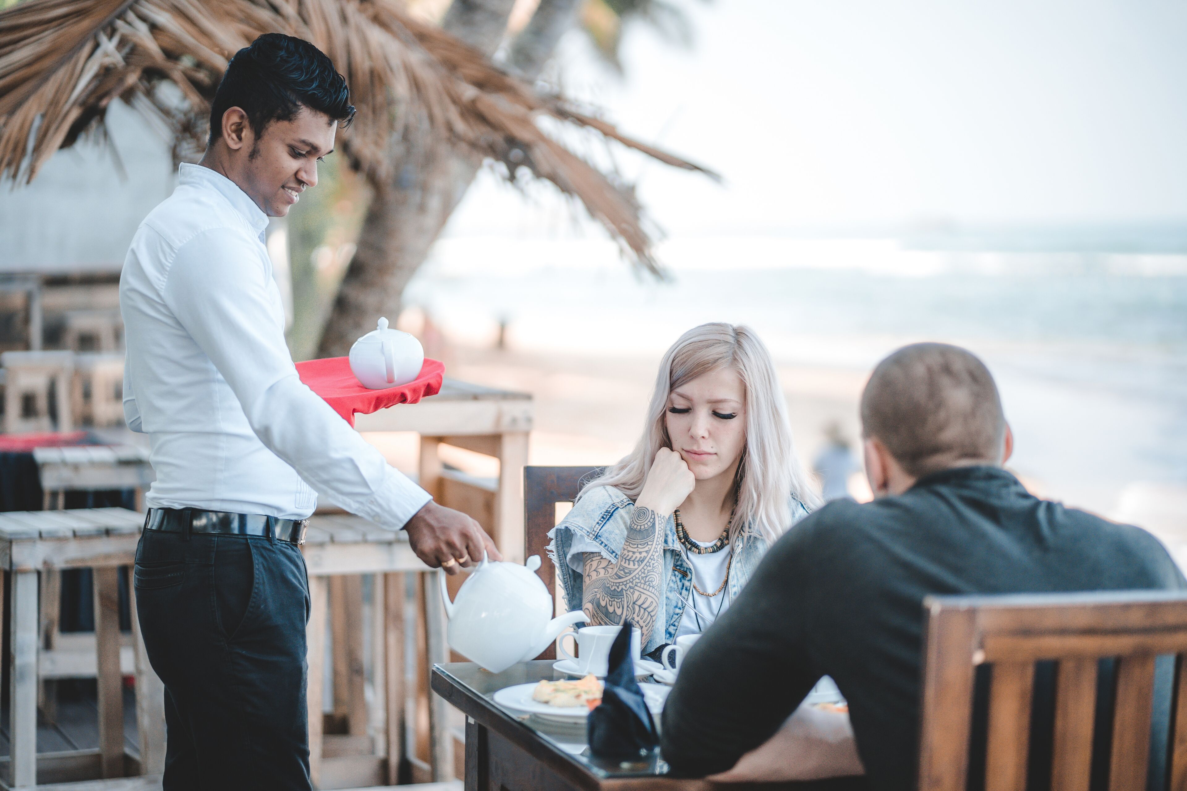A beachside café with wooden tables and chairs, a palm tree in the background, and a view of the ocean.