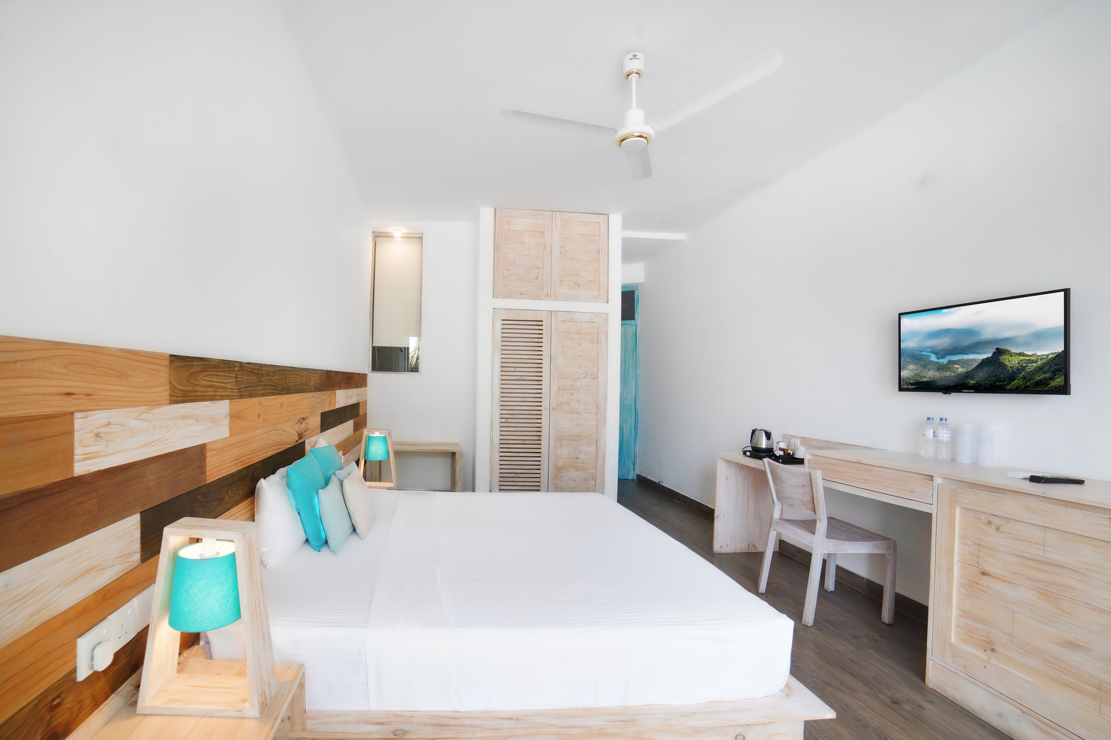 A modern bedroom with a wooden headboard, a bed with white bedding, a desk with a chair, a television, and a ceiling fan.