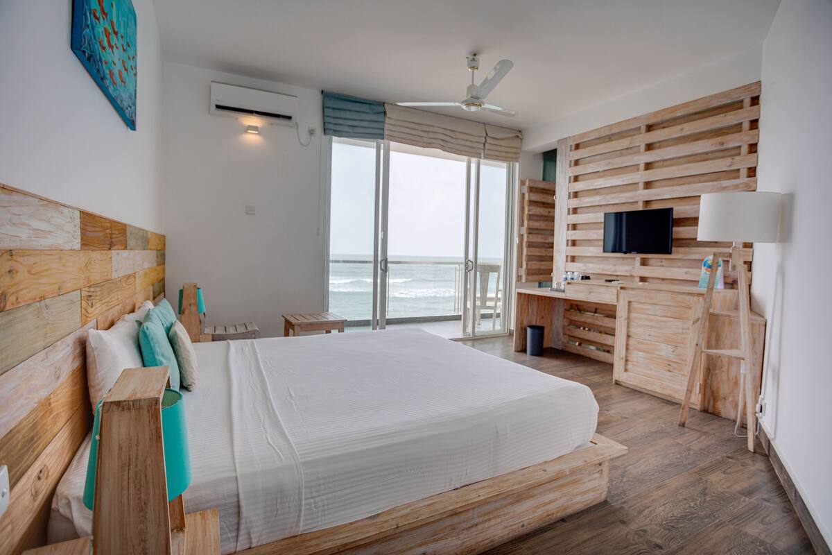 Une chambre avec un grand lit, une tête de lit en bois, un bureau avec une télévision et une vue sur l’océan.