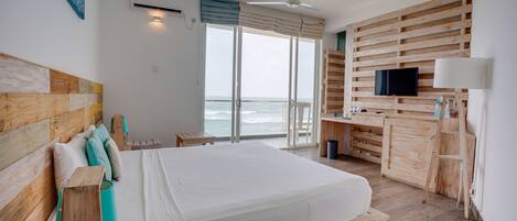 A bedroom with a large bed, wooden headboard, a desk with a TV, and a view of the ocean.