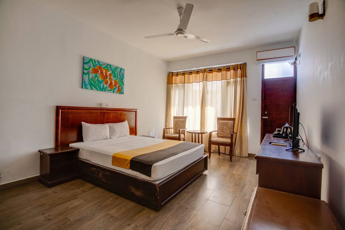 Une chambre d’hôtel avec un lit en bois, un ventilateur de plafond, un bureau avec un ordinateur et une fenêtre avec des rideaux.