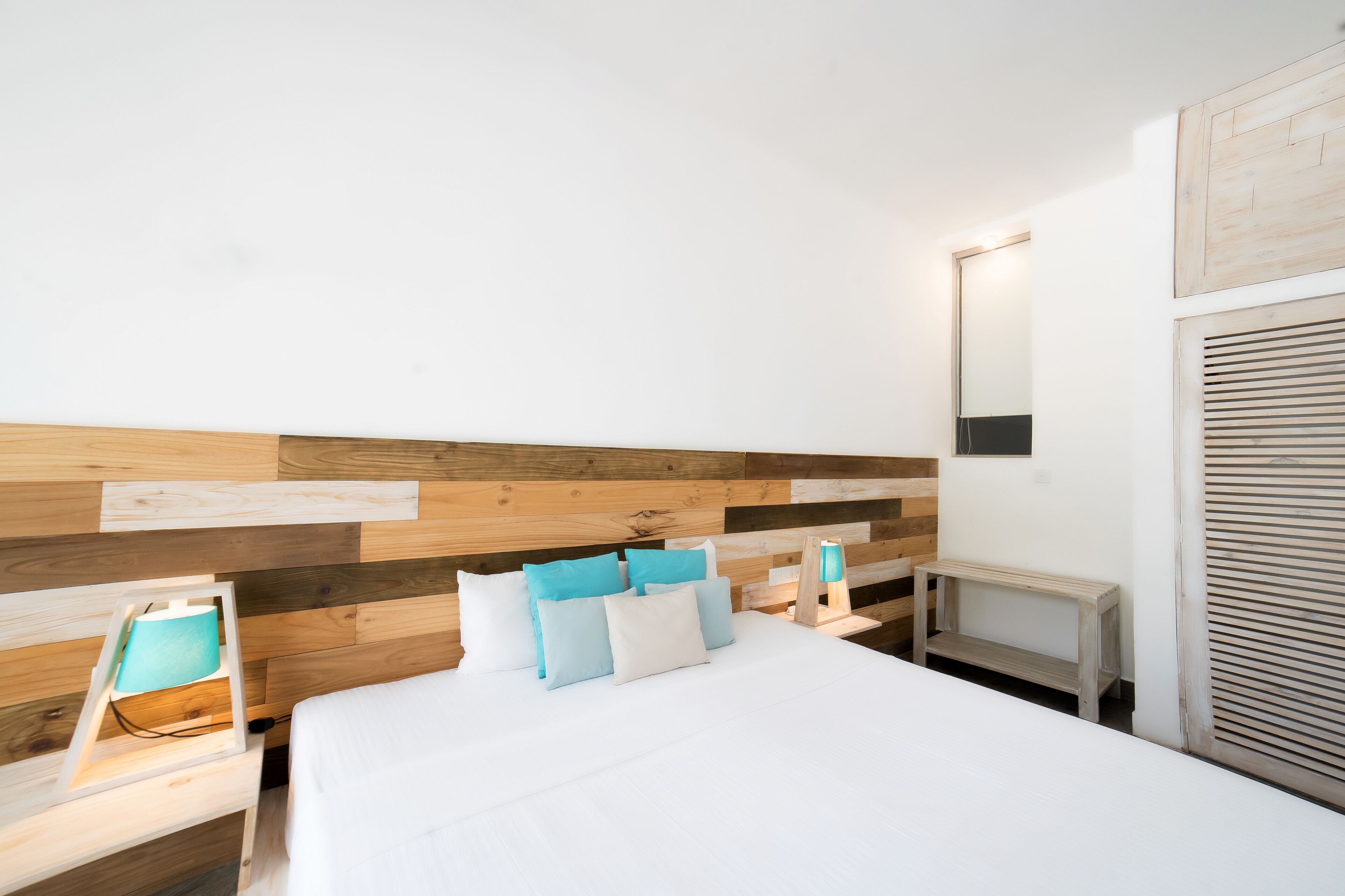 A bedroom with a wooden headboard, a bed with white and blue bedding, a bedside table with a lamp, and a small wooden bench.