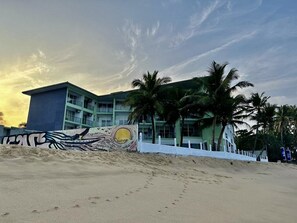 Et strandhotel med moderne design, der viser en stor bygning med flere balkoner, palmer og en sandstrand.