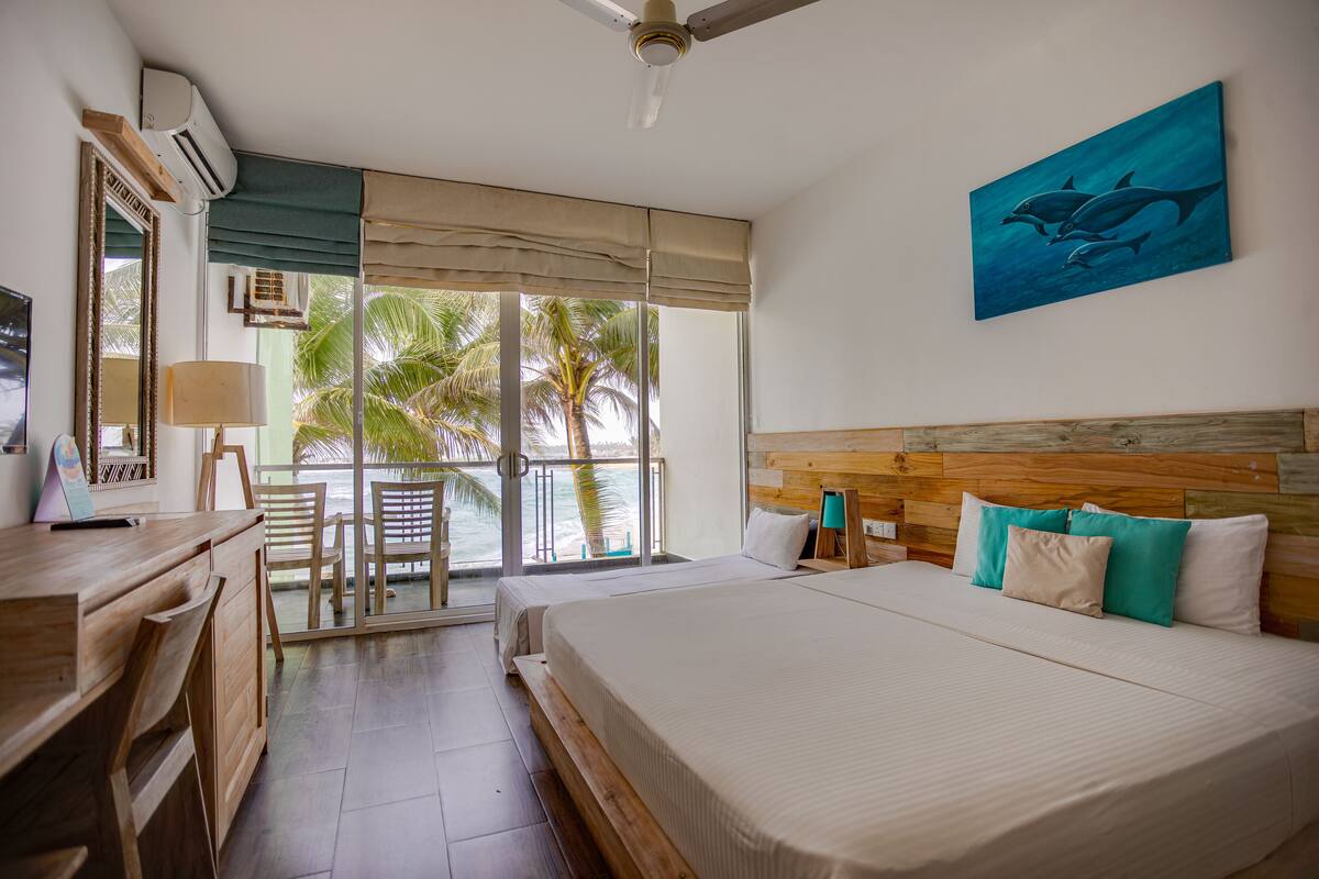 Une chambre d’hôtel avec un lit, un bureau, une chaise, un balcon donnant sur des palmiers et un tableau représentant des dauphins accroché au mur.