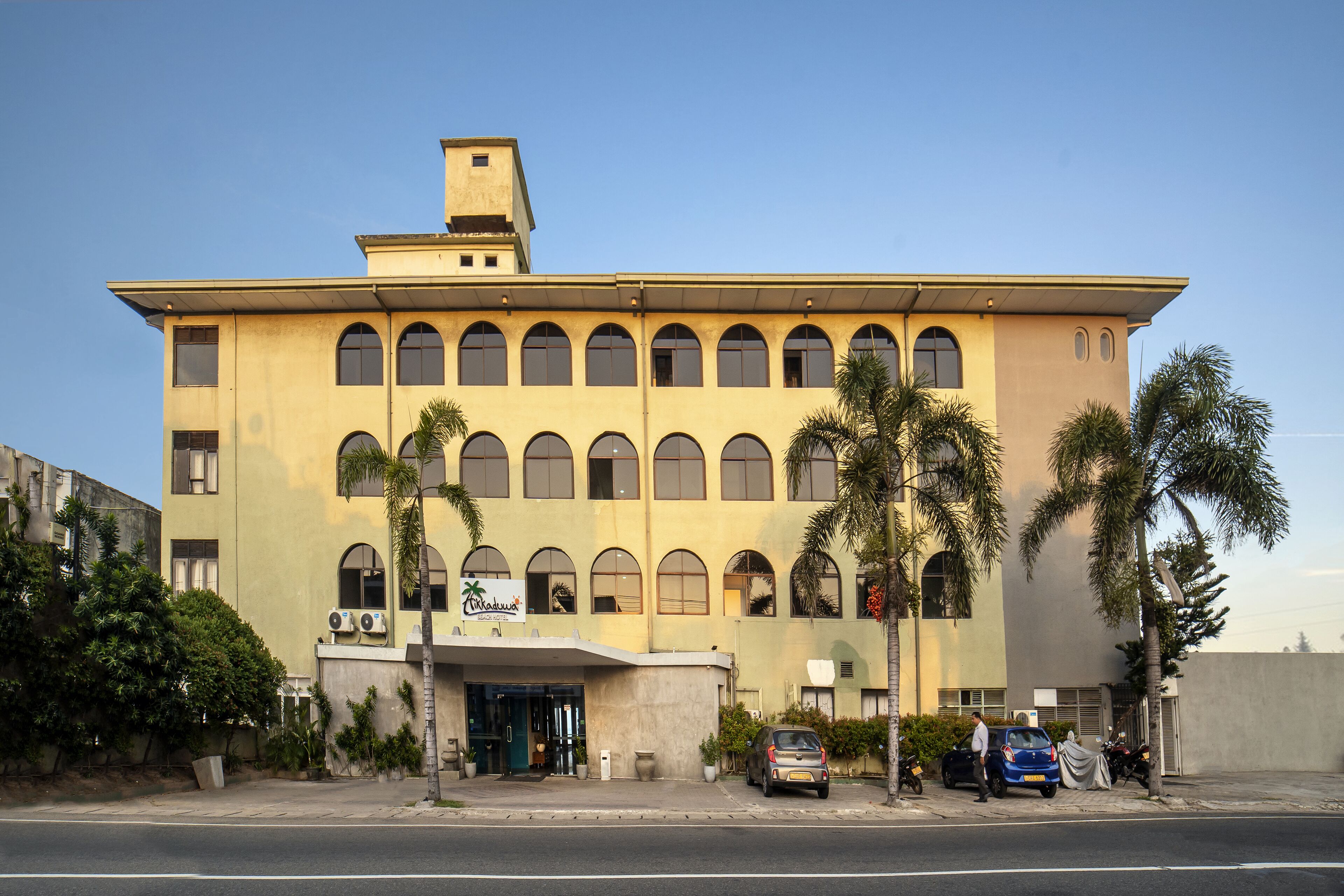 Un edificio di due piani con tetto piatto, numerose finestre ad arco e una insegna che indica che si tratta di un hotel. Palme si ergono davanti e alcune auto sono parcheggiate all'esterno.