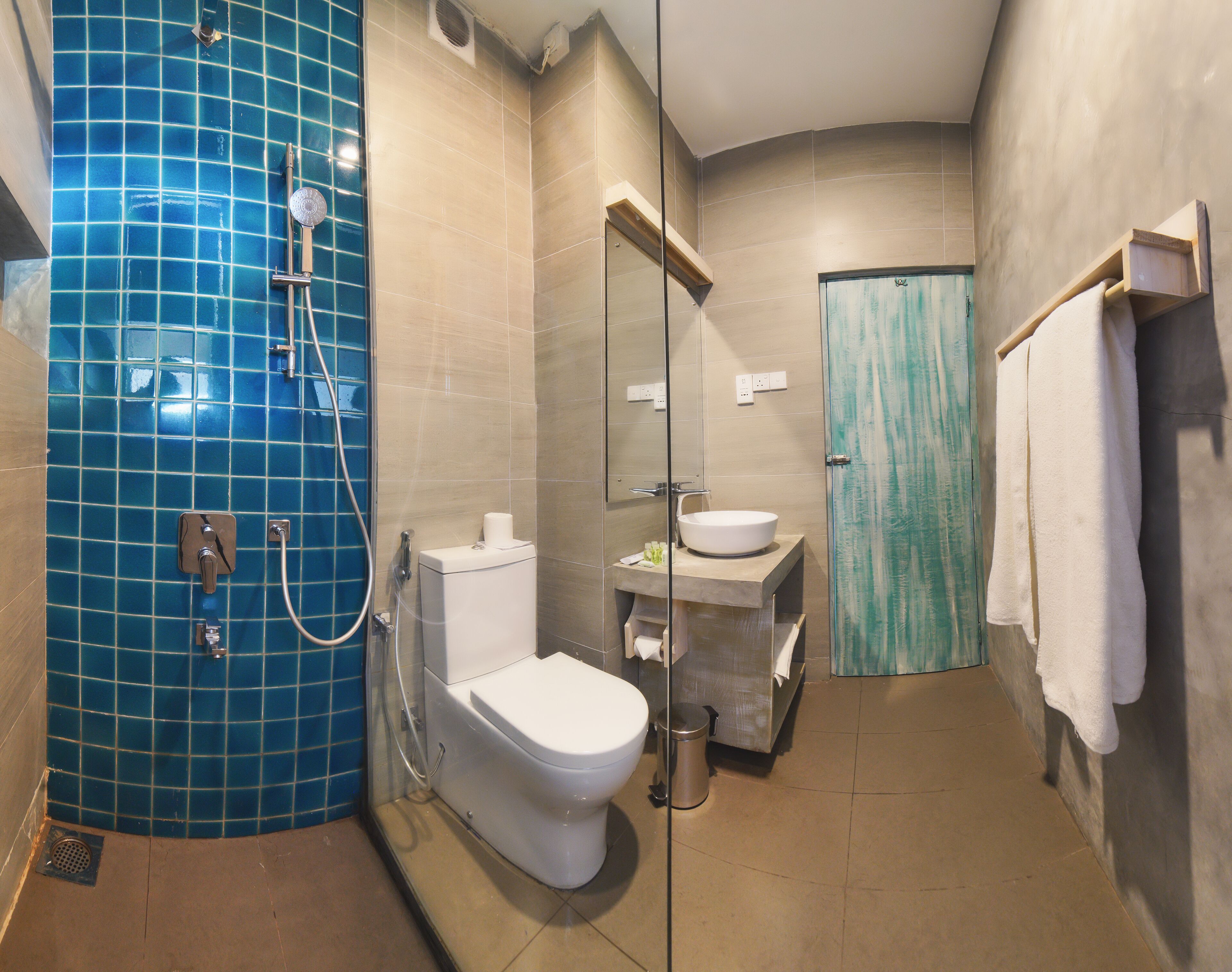 Une salle de bain avec un mur de douche recouvert de carreaux bleus, des toilettes blanches, un lavabo avec un miroir et un porte-serviettes.