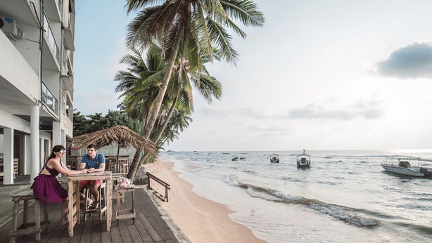 Un patio frente a la playa con mesa y sillas de madera, con vista al océano, barcos y palmeras.