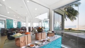 Un comedor moderno con una barra de buffet, grandes ventanales con vista a la playa y una planta tropical en la esquina.