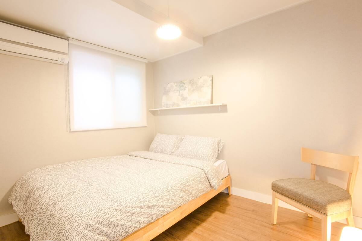 A bedroom with a bed, a chair, a window with blinds, a wall-mounted shelf, and an air conditioning unit.
