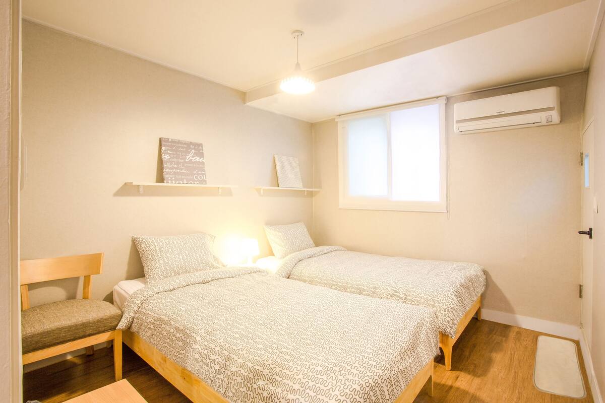 A bedroom with two single beds, a wooden chair, a bedside table, a wall-mounted air conditioner, and a window with blinds.