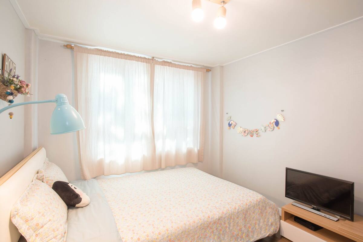 A bedroom with a bed, a TV on a stand, a lamp, and a window with curtains.