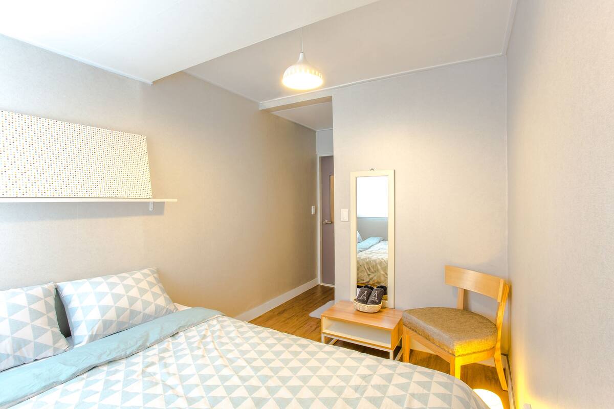 A bedroom with a bed, a chair, a small table, a mirror, and a hanging light fixture.