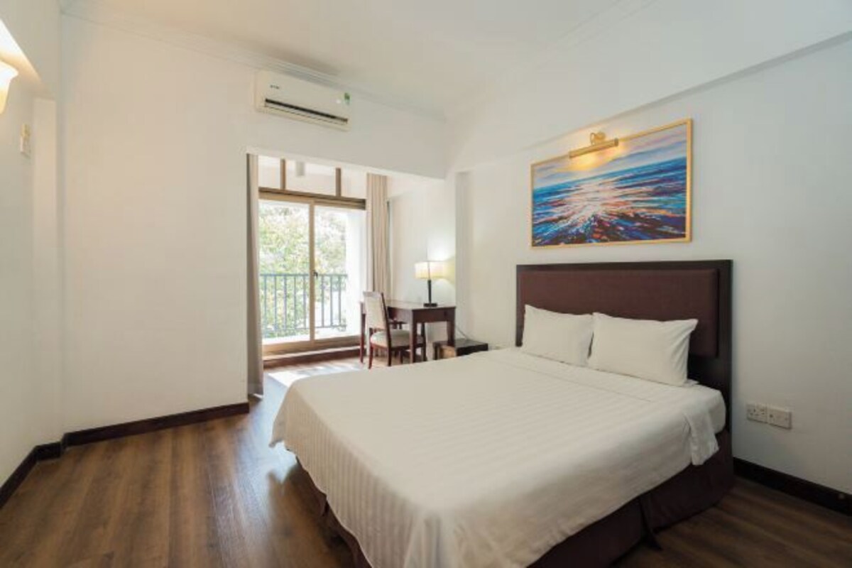 A hotel room with a large bed, a desk with a chair, a window with a view, and a painting on the wall.