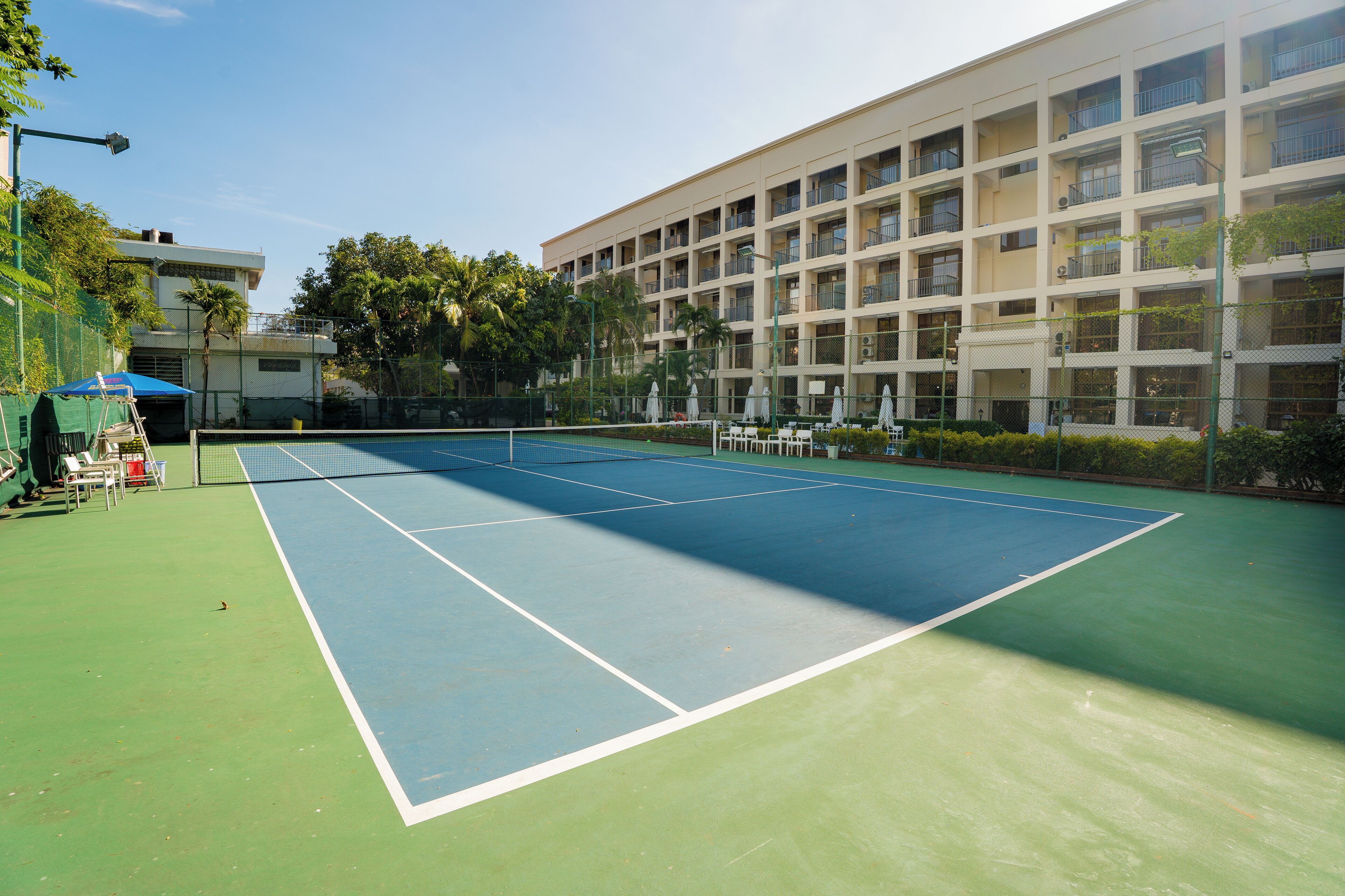 A tennis court with a net, surrounded by a fence, and adjacent to a building with multiple balconies.