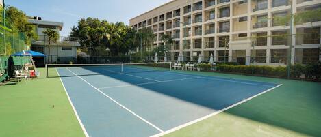 A tennis court with a net, surrounded by a fence, and adjacent to a building with multiple balconies.