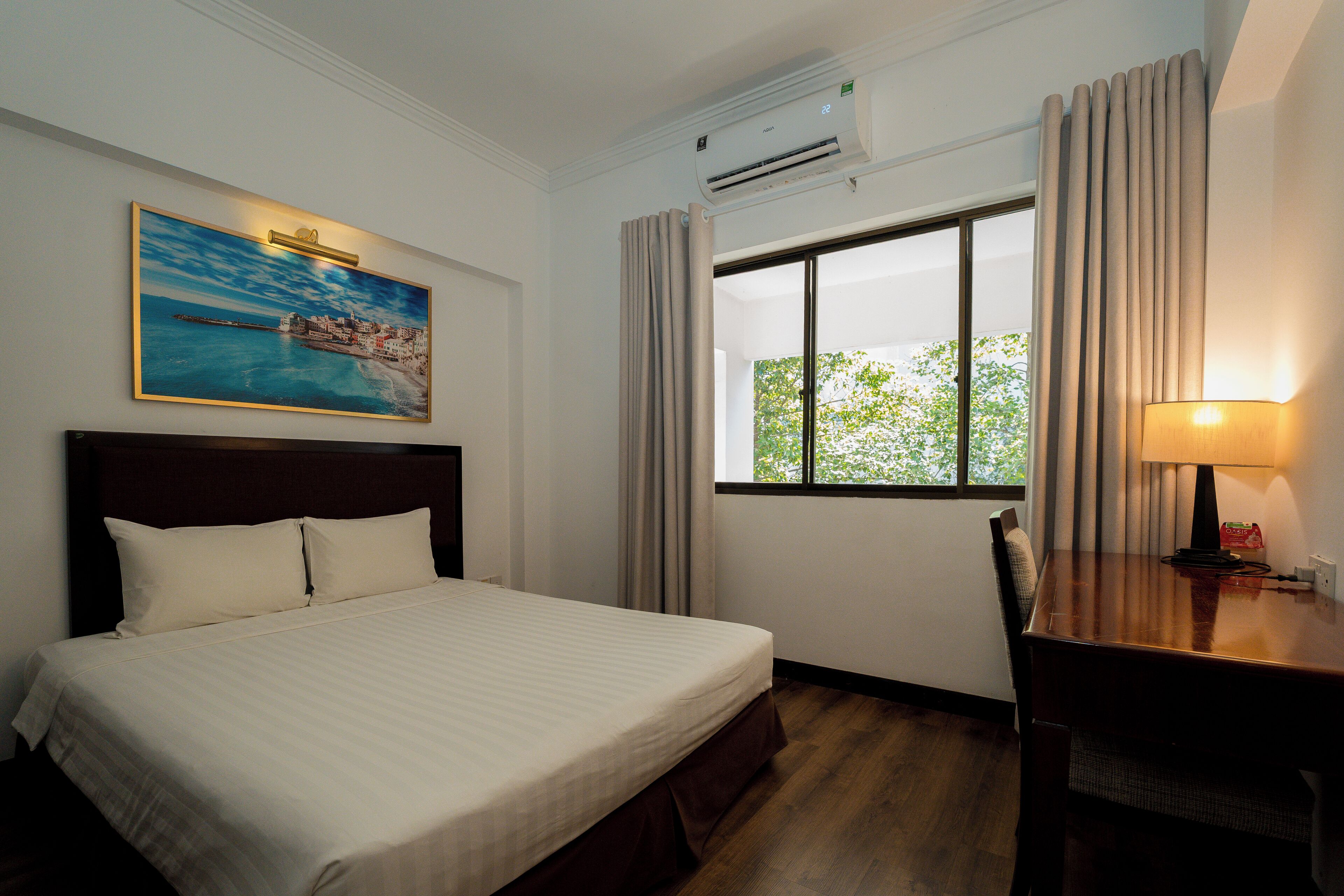 A hotel room with a bed, a desk, a lamp, an air conditioner, a window with curtains, and a painting on the wall.