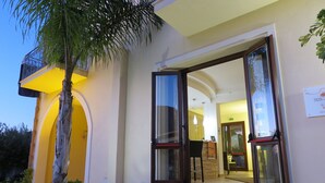 Entrada de hotel com portas abertas que dão para uma área de recepção, uma palmeira e uma escadaria.