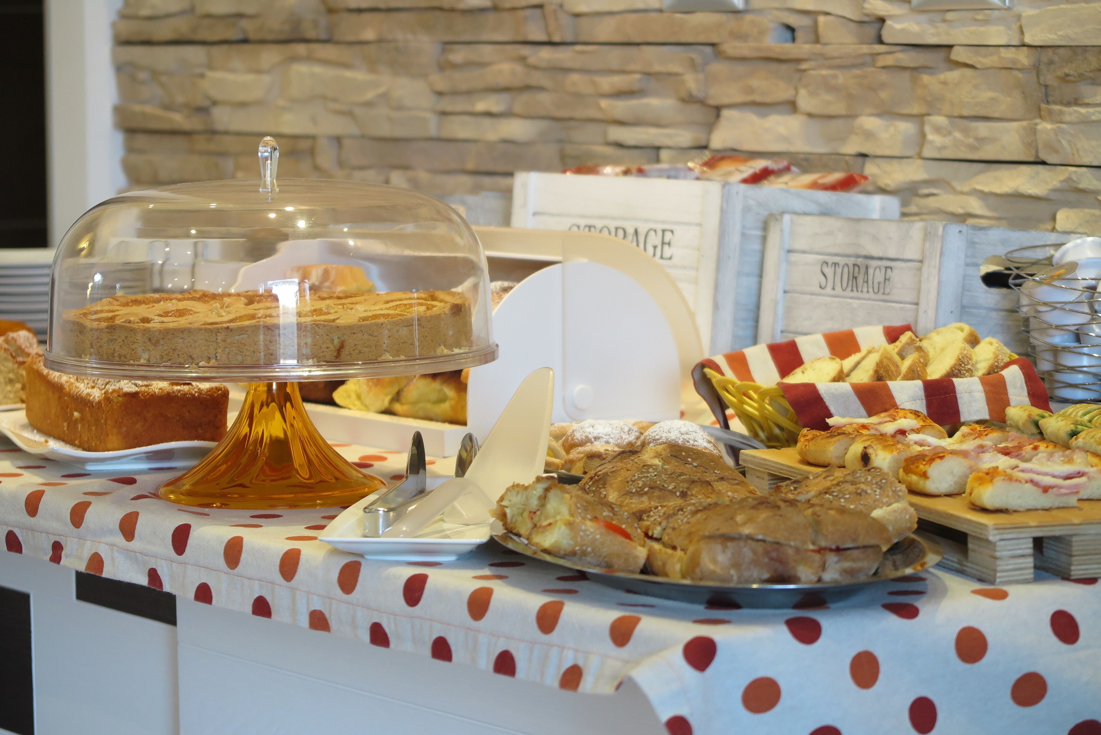 Un tavolo per buffet con diversi cibi, tra cui una torta coperta, panini e pasticceria, su sfondo di una parete in pietra.