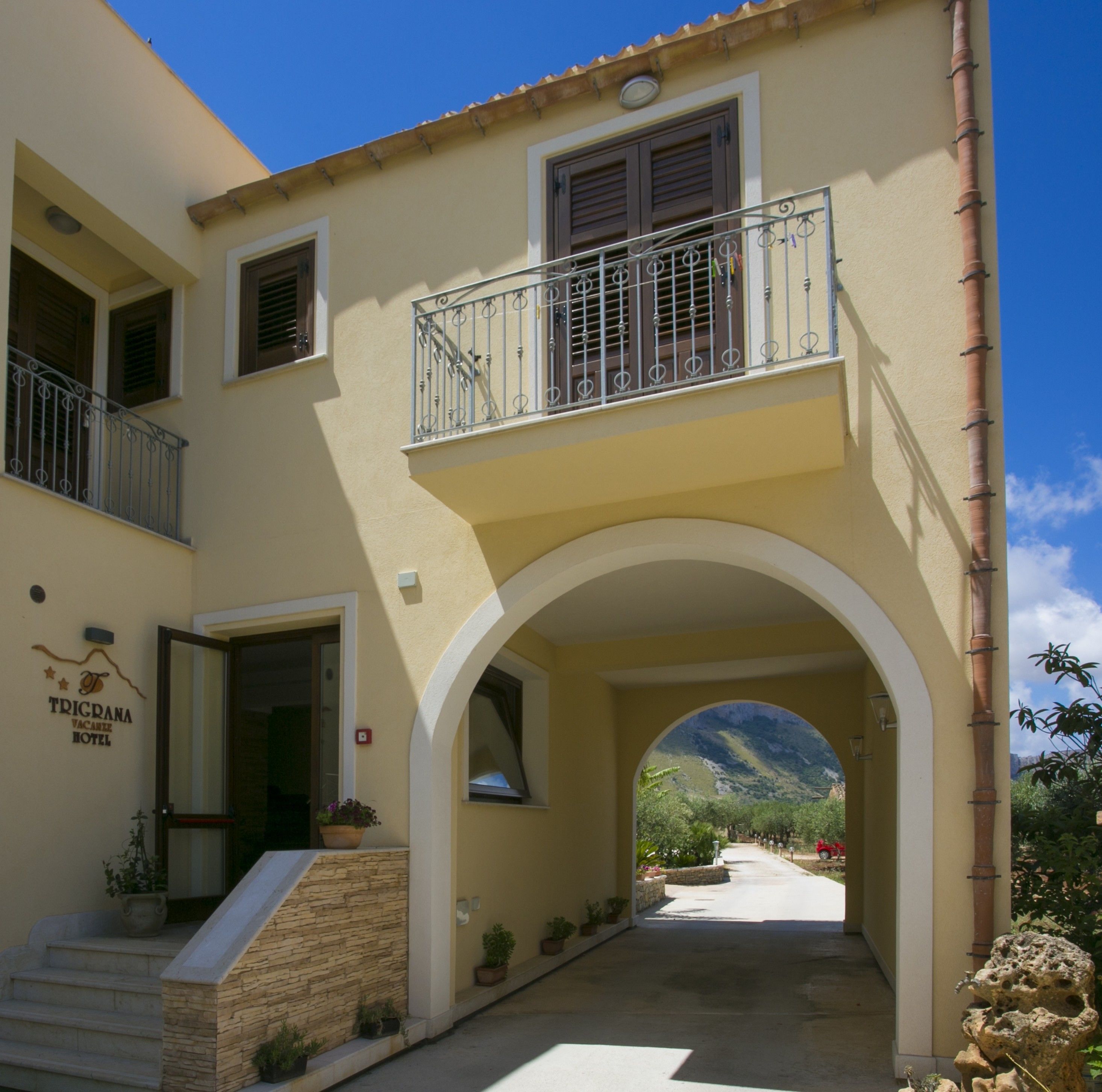 Un edificio a due piani con un balcone, un arco che offre vista sulle montagne e un cartello per l'Hotel Trigrana Vacanze.
