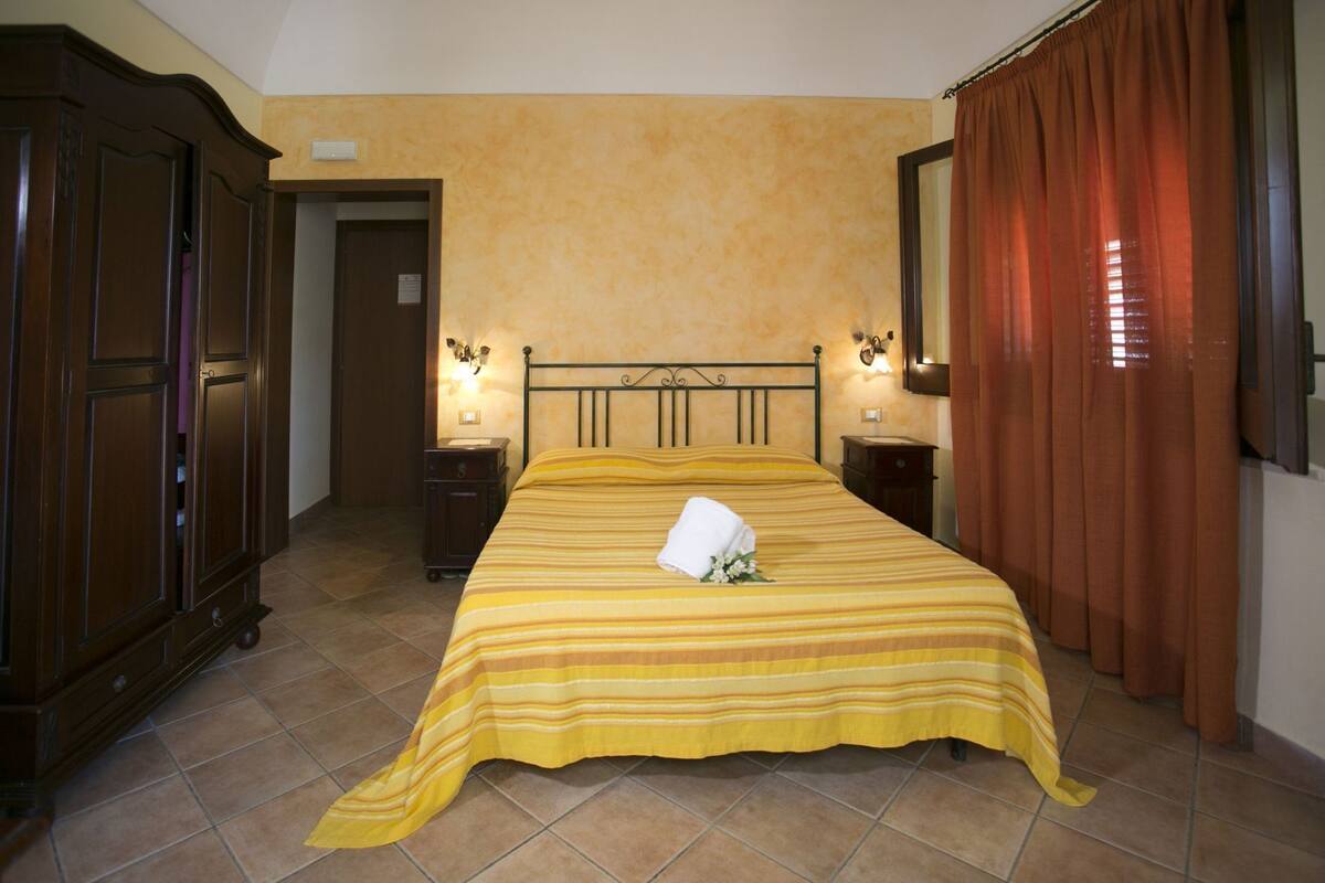 Une chambre d’hôtel comprenant un lit, une armoire, une table de chevet, un miroir et une fenêtre avec des rideaux.
