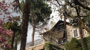 Un edificio de piedra con balcón, rodeado de árboles y arbustos.