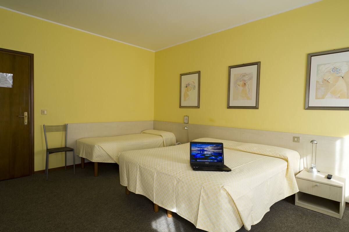 Quarto de hotel com duas camas de solteiro, uma escrivaninha com um laptop, uma cadeira e quadros nas paredes.
