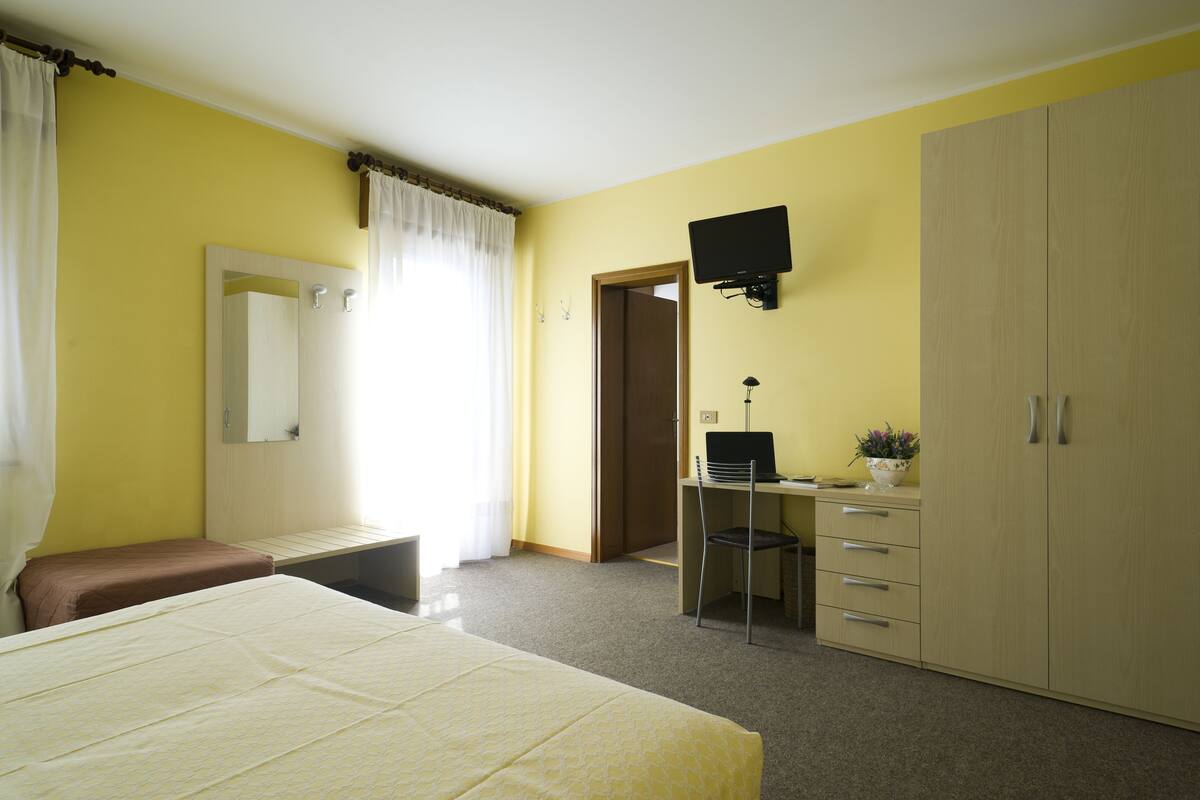 Quarto de hotel com uma cama, uma escrivaninha com cadeira, um guarda-roupa e uma televisão na parede.