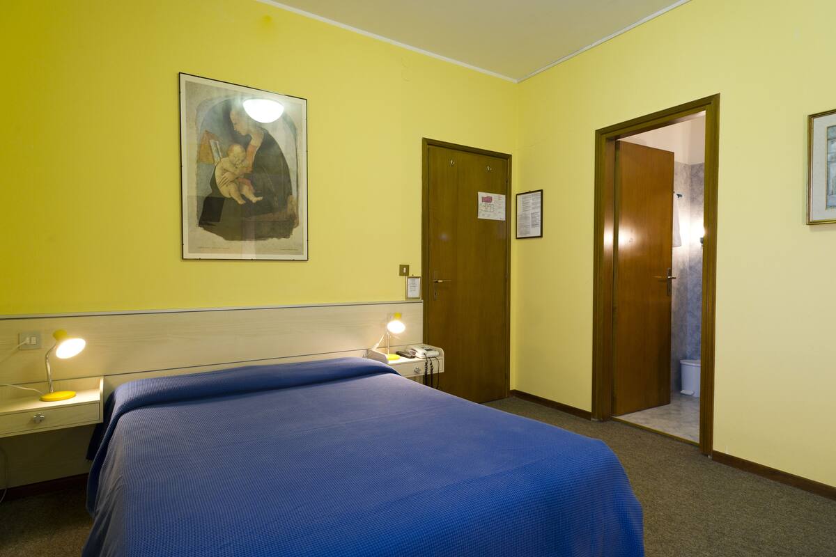 Quarto de hotel com cama, mesa de cabeceira, luminária e porta para um banheiro.