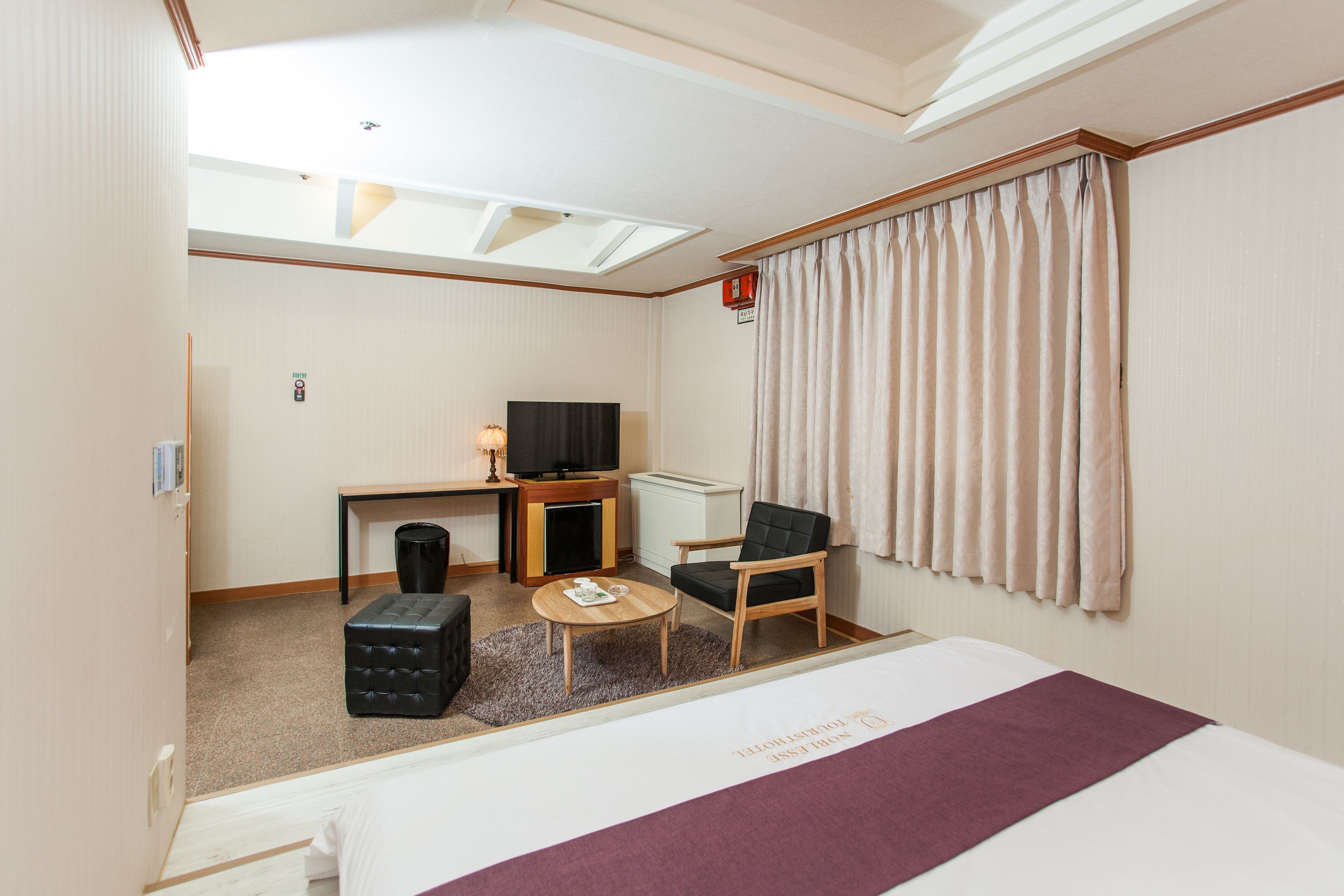 Una habitación de hotel con una cama, un escritorio con televisor, una silla, una mesita y una ventana con cortinas.