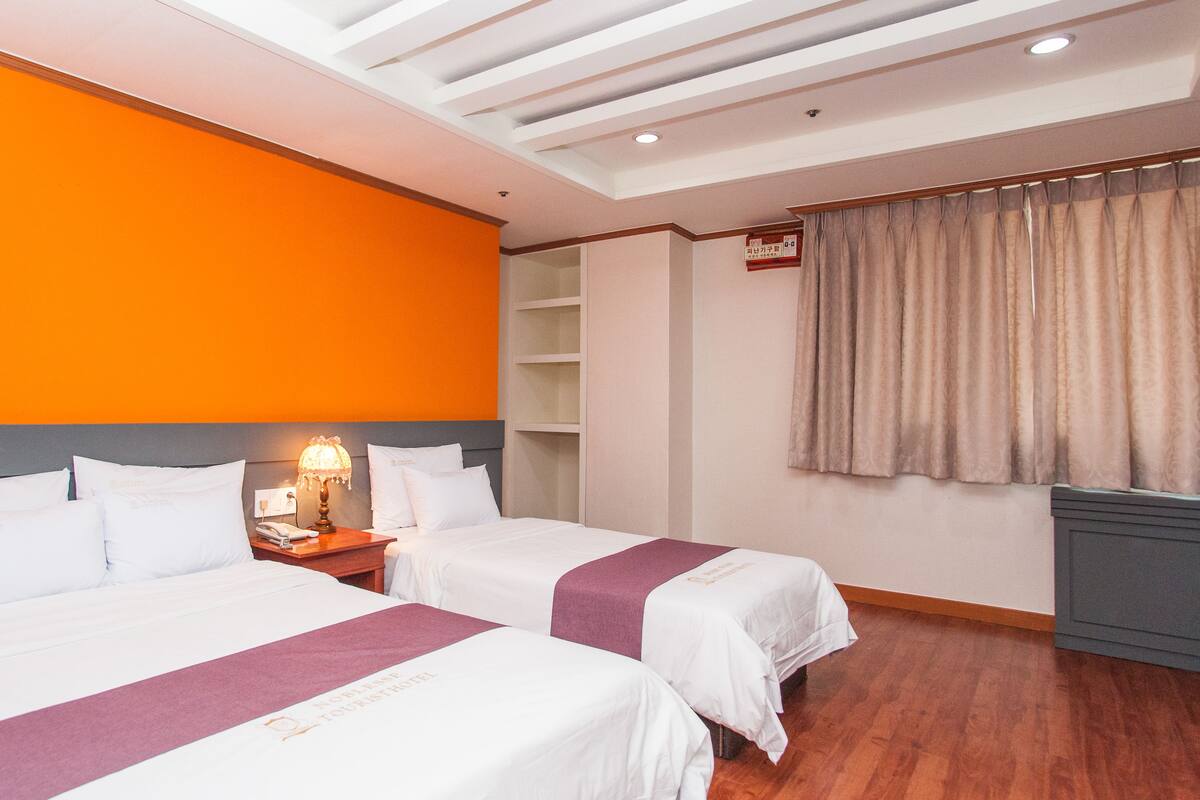 A hotel room with two beds, a wooden floor, an orange accent wall, a nightstand with a lamp, and a window with curtains.