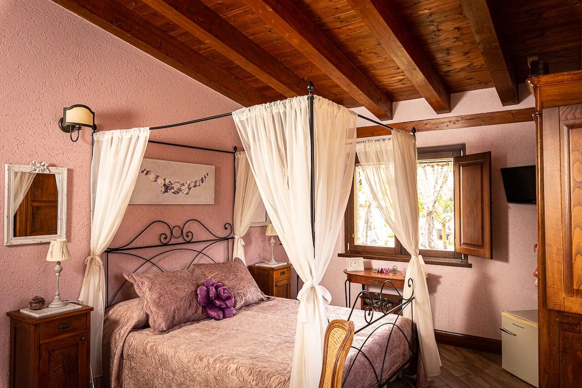 A bedroom with a four-poster bed, a nightstand, a desk, and a small refrigerator.