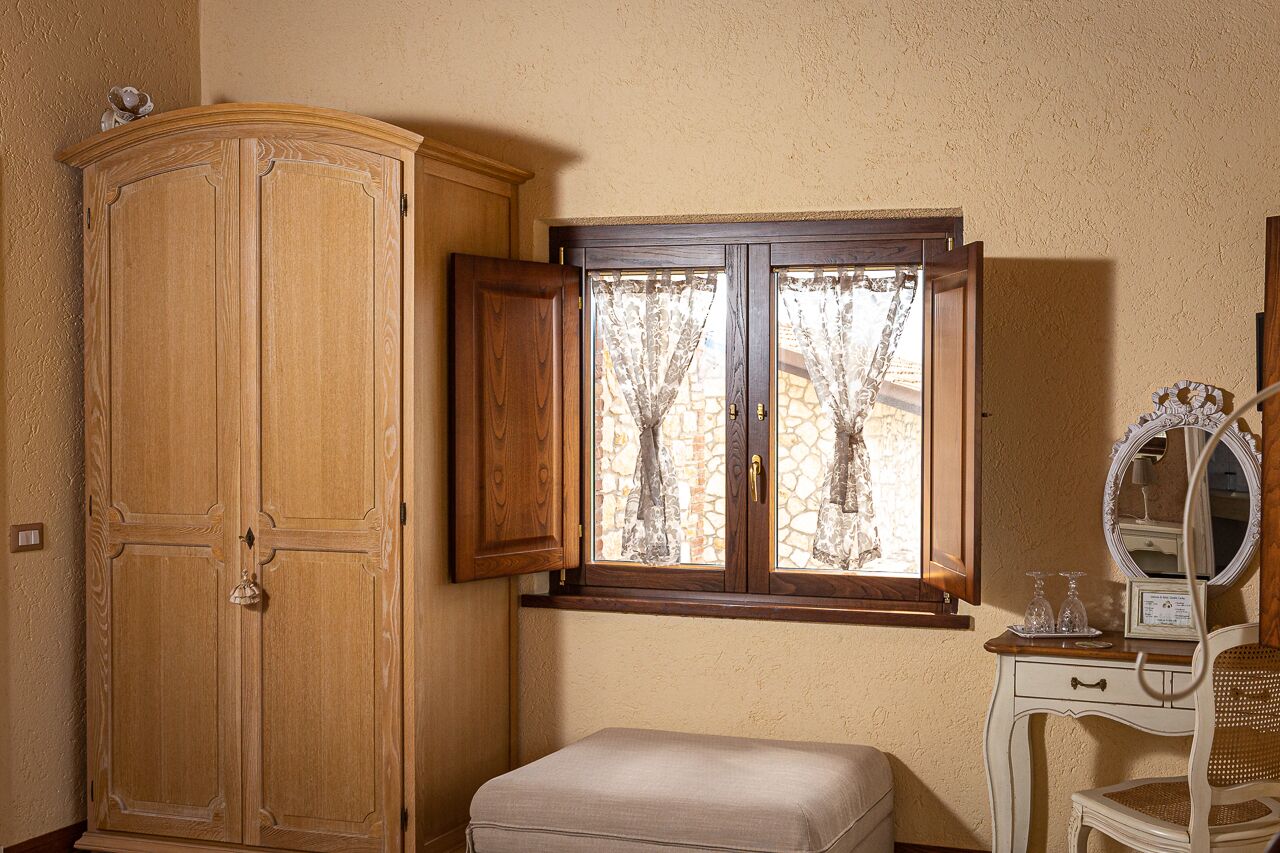 A room with a wooden wardrobe, a window with curtains, a vanity with a mirror, and a bench.