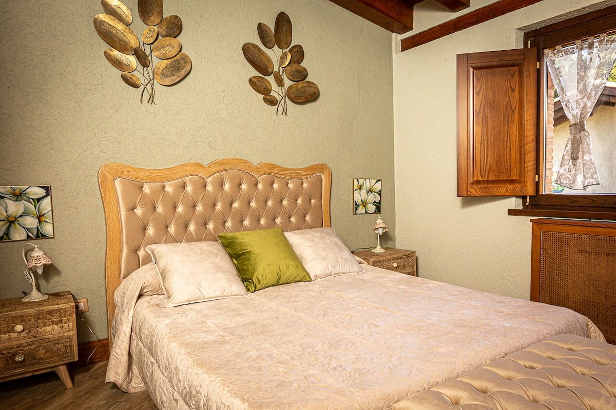 A bedroom with a tufted headboard, a bed with a patterned bedspread, two wall-mounted decorative leaf motifs, a wooden nightstand with a lamp, and a window with a wooden frame and lace curtains.