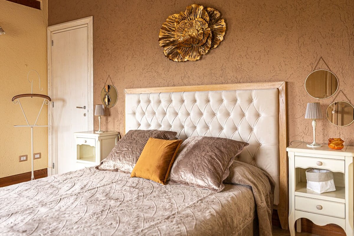 A bedroom with a tufted headboard, a patterned bedspread, two pillows, a bedside table with a lamp, and a wall decoration.