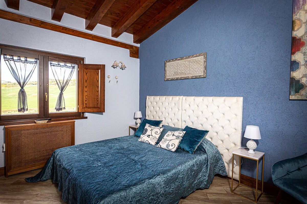 A bedroom with a large bed, a wooden headboard, a window with curtains, a wall-mounted lamp, and a small table.
