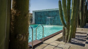 Una piscina con cascada, rodeada de cactus y un muro de piedra.