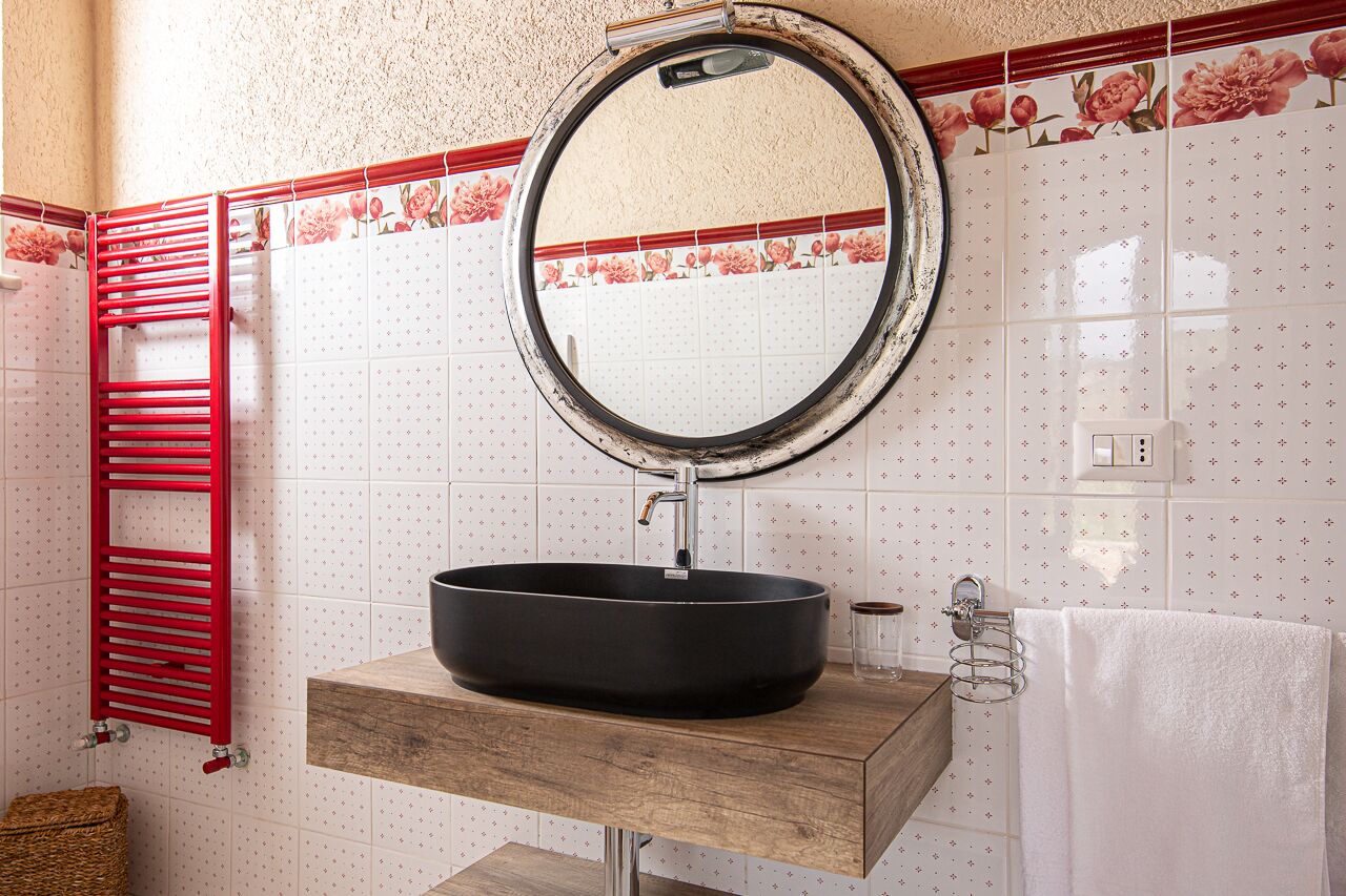 Ein Badezimmer mit rundem Spiegel, schwarzem Waschbecken, Holz-Waschtisch, rotem Handtuchhalter und Fliesen mit Blumenmuster.
