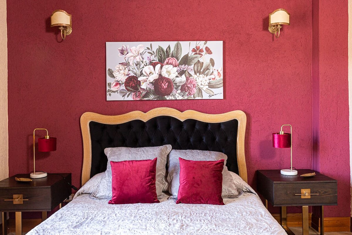 A bedroom with a tufted headboard, a floral artwork on the wall, two bedside tables with lamps, and two red pillows on the bed.