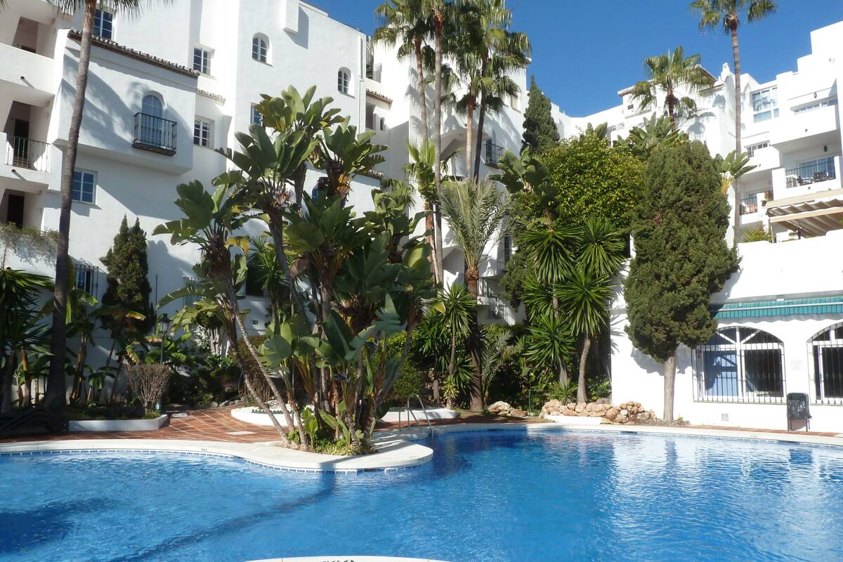 A swimming pool surrounded by white buildings, palm trees, and a clear blue sky.