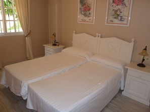 Una cama doble con cabecera blanca, una mesita de noche con lámpara y otro pequeño mueble con lámpara. En la pared, dos cuadros enmarcados con motivos florales.