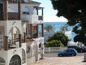 A coastal building with multiple balconies, a visible sea view, and a paved street with parked cars.
