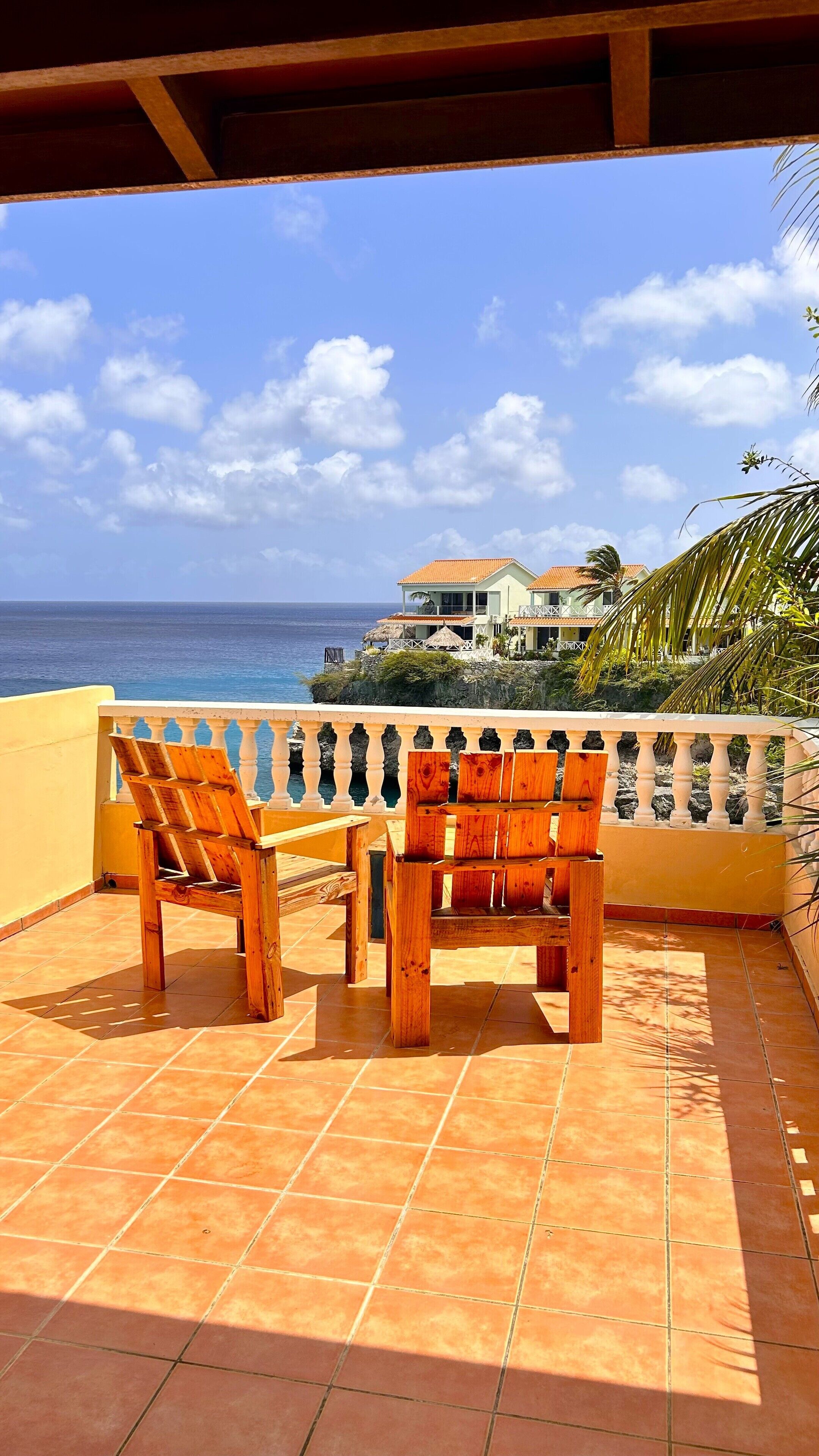 Una terraza con sillas y una mesa de madera, con vistas al mar y una casa al fondo.