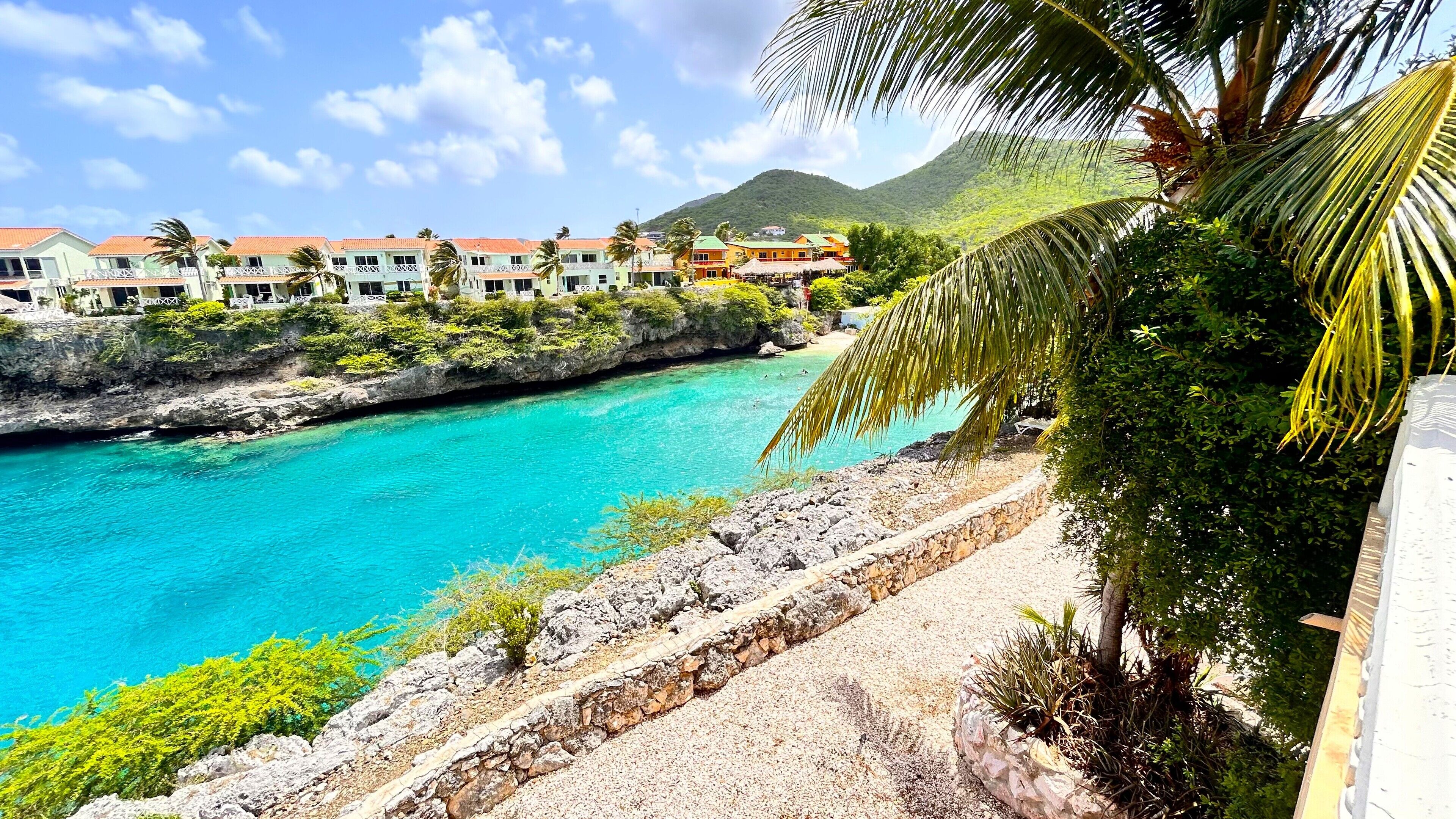 A coastal hotel with a turquoise sea view, stone pathway, and lush greenery.