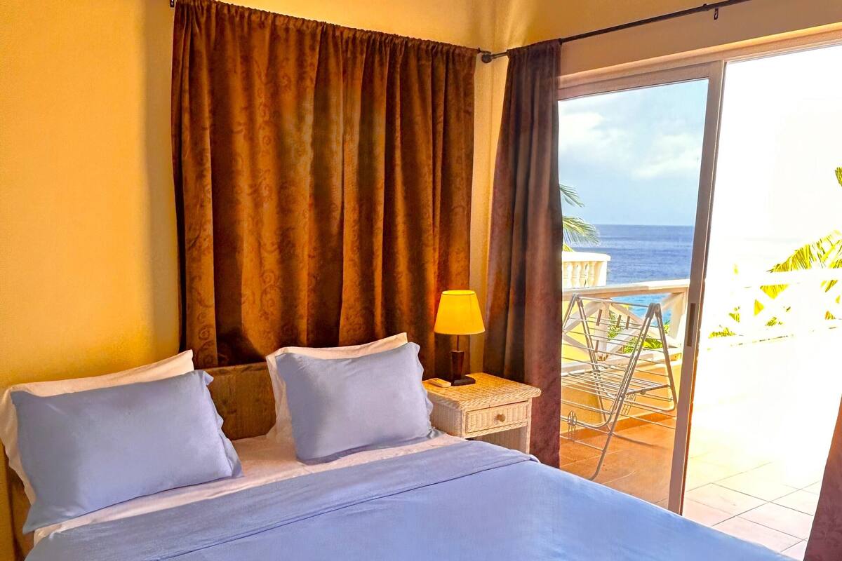 Habitación de hotel con cama, mesita de noche, lámpara y vista al océano.