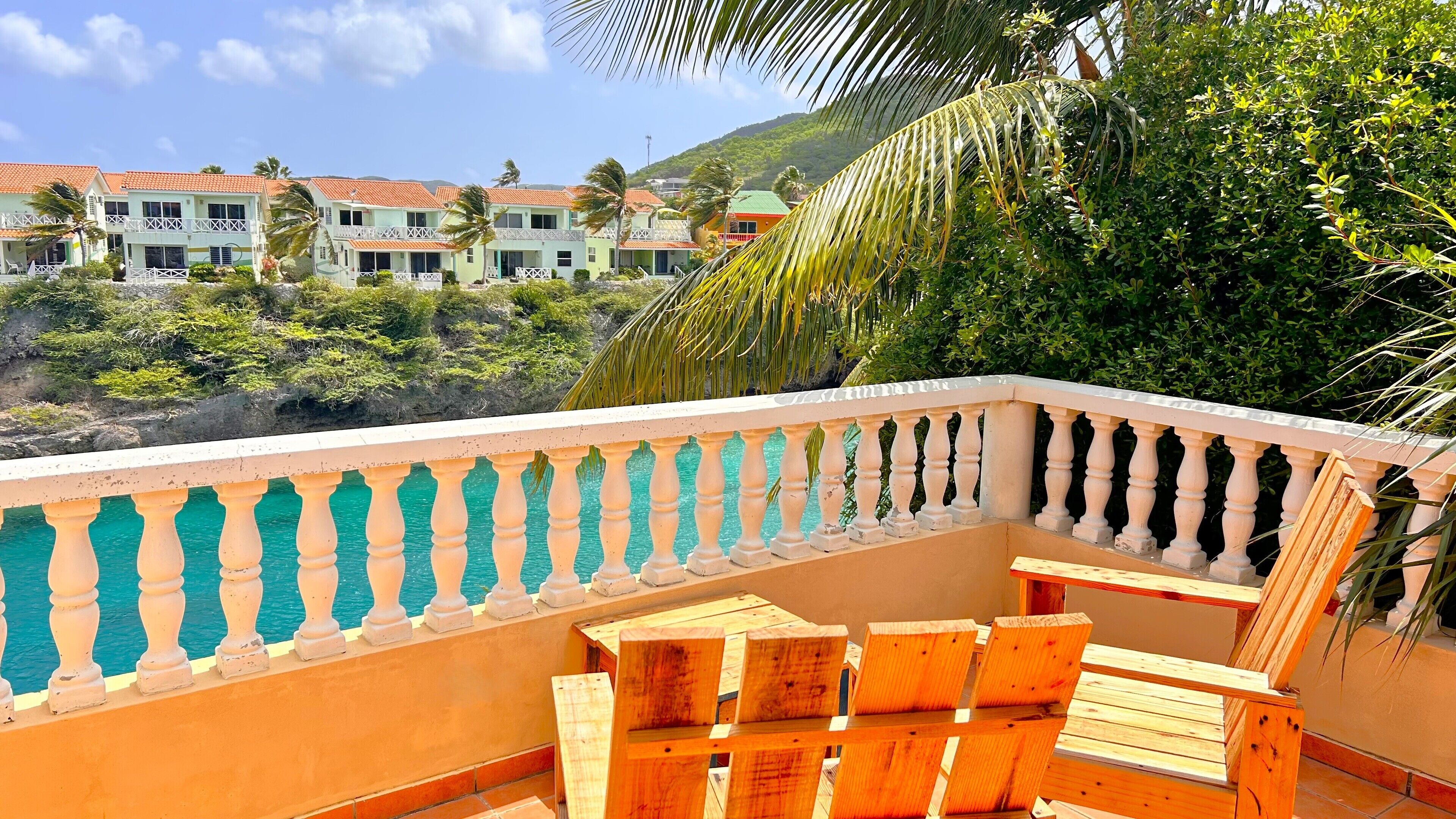 A balcony with wooden chairs and a view of a resort with multiple buildings and palm trees.