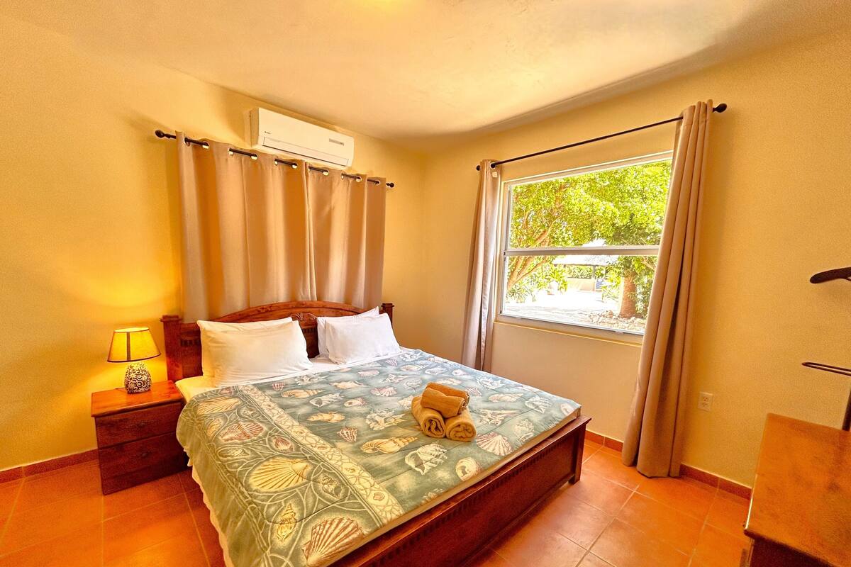 Habitación de hotel con cama, mesita de noche con lámpara, equipo de aire acondicionado, ventana con cortinas y vista a zonas verdes.