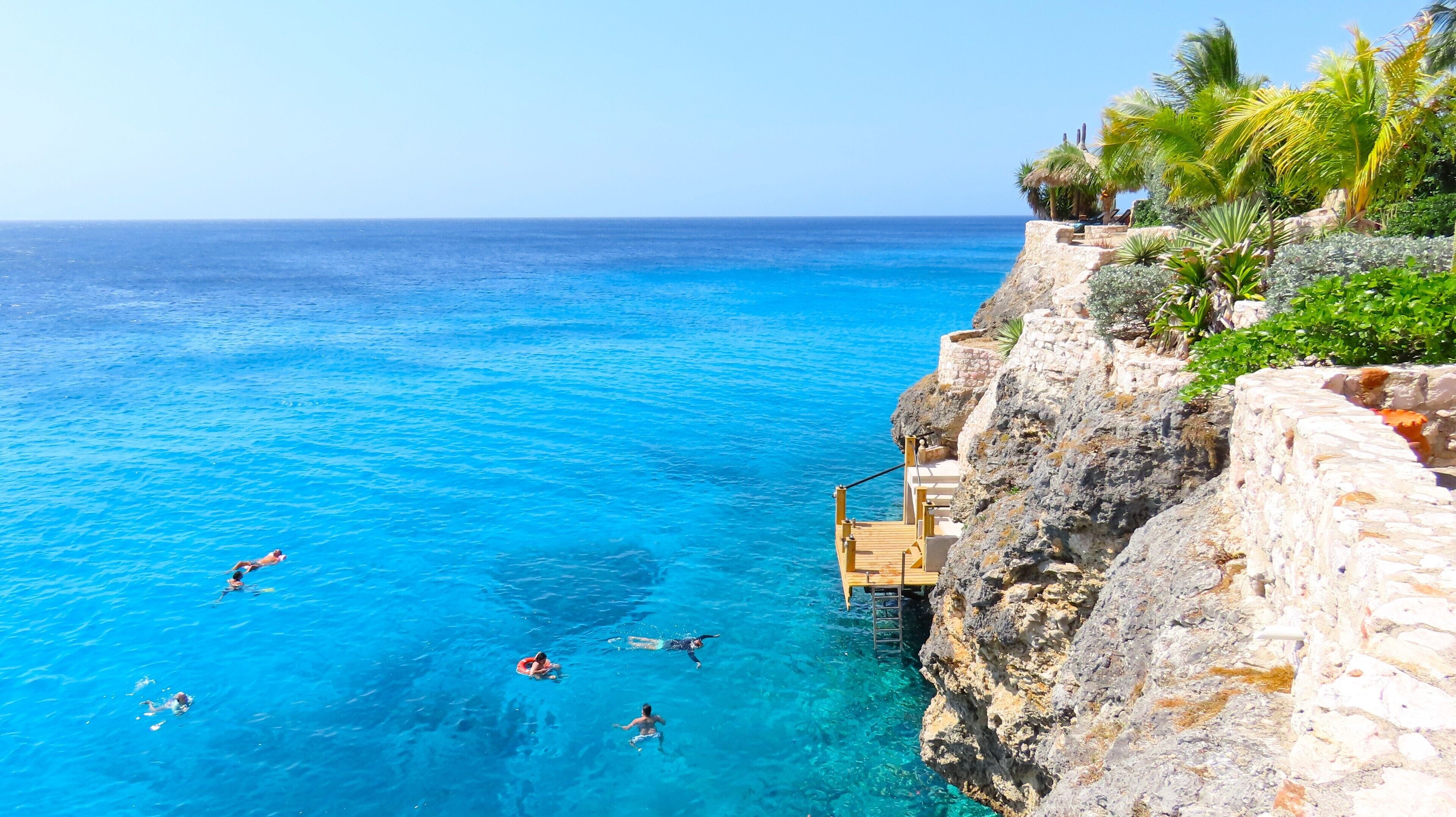 A coastal cliff with a wooden staircase leading down to clear blue water, where people are swimming and snorkeling.