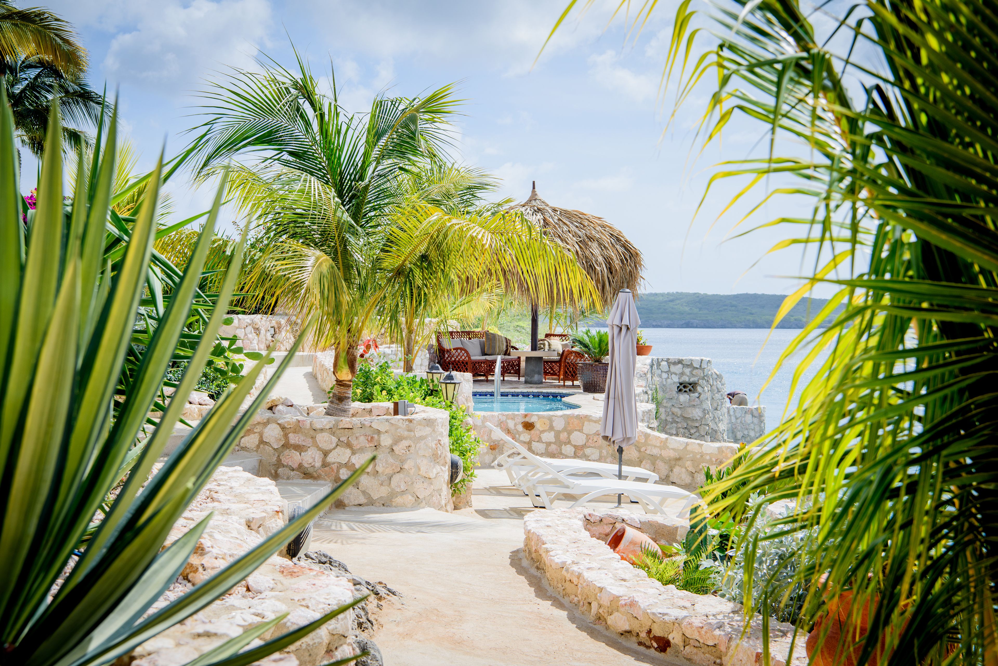 A pool area with lounge chairs, a thatched umbrella, and a stone wall.