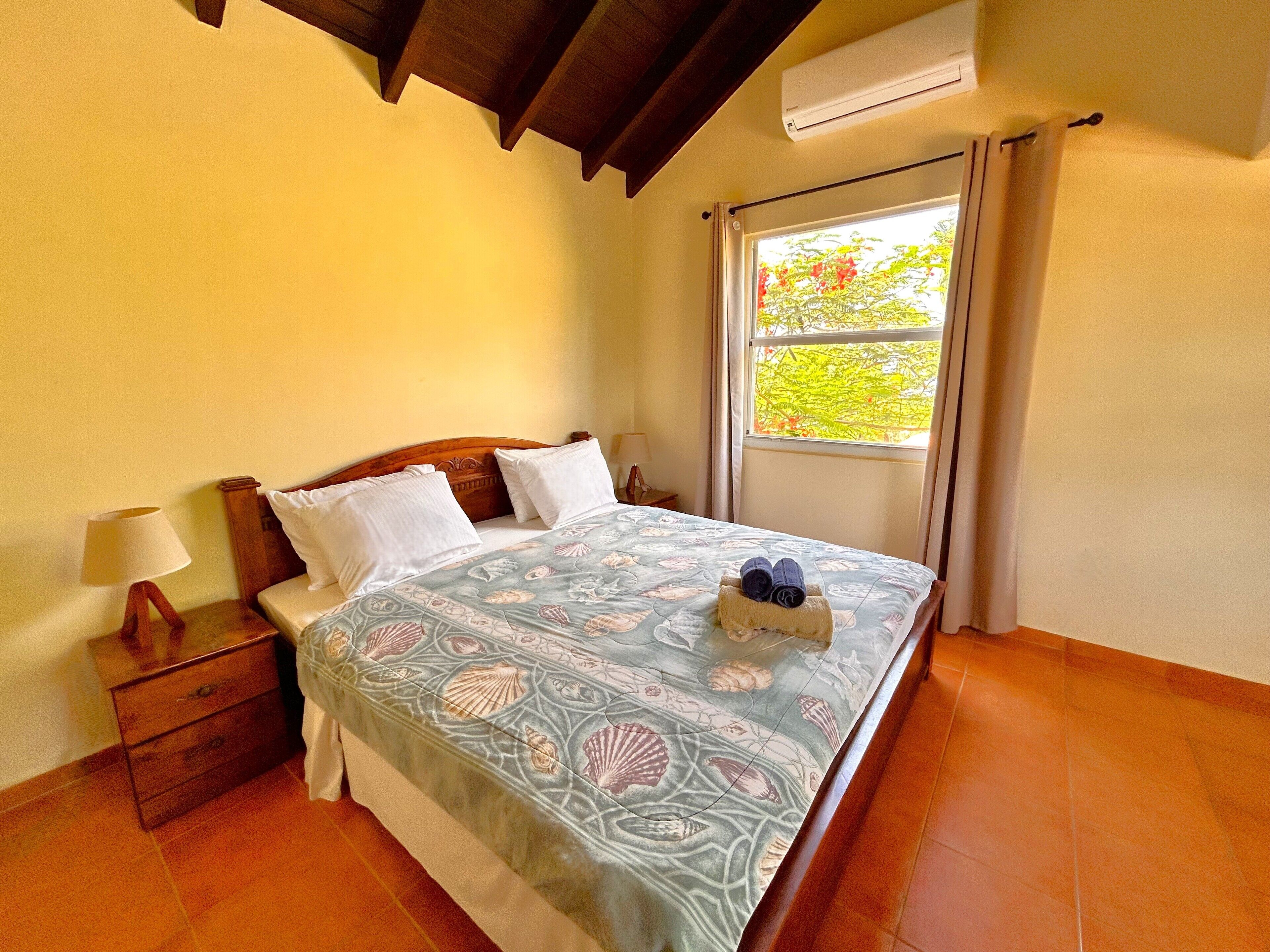 A neatly made bed with a floral bedspread, a nightstand with a lamp, and a window with curtains.